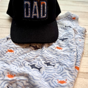 Pode incluir: Um boné de beisebol preto com a palavra "DAD" em um patch azul e branco. Ao lado do boné, um body de bebê azul claro e branco com um padrão de tubarões e caranguejos.