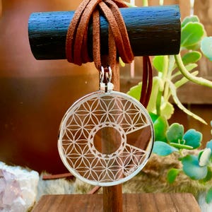 May include: A silver pendant with a geometric design, suspended from a brown leather cord. The pendant features a circular pattern with a cut-out section. The necklace is displayed on a wooden stand, with greenery in the background.