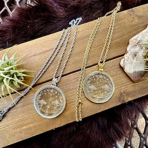 May include: Two pendant necklaces with round, clear glass pendants featuring a mushroom design, stars, and foliage. One necklace has a silver chain, the other a gold chain. The pendants are displayed on a wooden surface.
