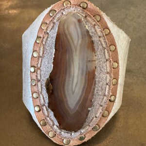 May include: A handcrafted leather cuff bracelet featuring a polished agate stone. The agate displays bands of brown, white, and cream. The bracelet has a light tan leather border with brass rivets and a white leather backing.