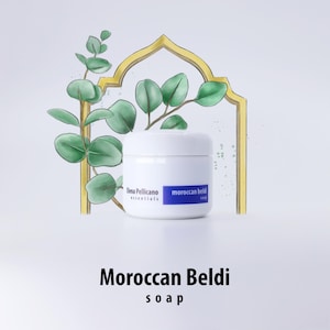 May include: A white jar of Moroccan Beldi soap with a blue label that reads "moroccan beldi soap" and "Bena Pellicano essentials". The jar is sitting on a white surface with a watercolor illustration of a eucalyptus branch in a gold frame.