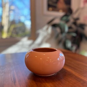 May include: A small, round, peach-colored ceramic vase with a glossy finish. The vase has a wide opening and sits on a wooden surface. The interior of the vase is a darker shade of peach. Home decor.