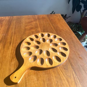 May include: A wooden serving tray with a handle, featuring numerous oval-shaped indentations. The tray is made of light-colored wood and is designed for serving appetizers or small portions. The handle has a small hole.