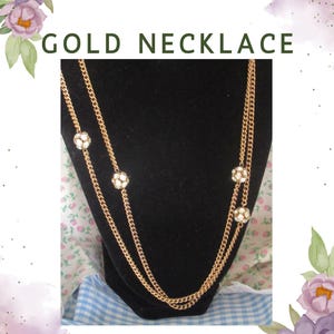 Può includere: Collana dorata con un design a doppia catena. La collana presenta tre decorazioni floreali decorative con cristalli trasparenti. Il testo "GOLD NECKLACE" è visualizzato nella parte superiore dell'immagine.
