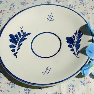 Könnte beinhalten: Ein weißer Keramikteller mit blauem Rand und blauen floralen Designs. Der Teller hat einen zentralen Kreis und zwei stilisierte Blattmuster. Der Teller liegt auf einer floral gemusterten Tischdecke.