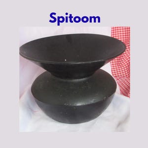 May include: A matte black spittoon featuring a wide, flared rim and a rounded, bulbous base. The word "Spitoom" is printed in blue text above the object. The vessel has a smooth, solid-colored finish and a distinct, symmetrical shape.