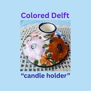 Puede incluir: Un portavelas de cerámica con un diseño floral. La base es redonda con una copa central para una vela. Las flores son en tonos de rosa, naranja y blanco, con hojas verdes. El texto "Colored Delft" y "candle holder" son visibles.
