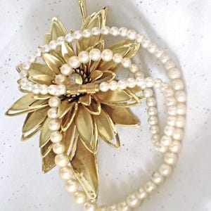 May include: A gold-toned metal flower brooch with layered petals and a central iridescent stone, layered with a single strand of white pearls featuring a cylindrical gold-toned clasp. The brooch is positioned behind the pearl necklace.