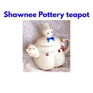 Puede incluir: Una tetera de cerámica Shawnee Pottery de color crema con una tapa en forma de payaso con pajarita azul y un pico en forma de cerdo. El cuerpo tiene pequeñas manchas marrones y el texto Shawnee Pottery teapot en la parte superior.
