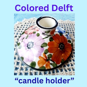 Può includere: Un portacandele rotondo in ceramica con un motivo floreale dipinto a mano caratterizzato da grandi fiori arancioni e viola chiaro, foglie verdi e un bordo blu scuro. Il testo "Colored Delft" appare in alto e "candle holder" in basso.
