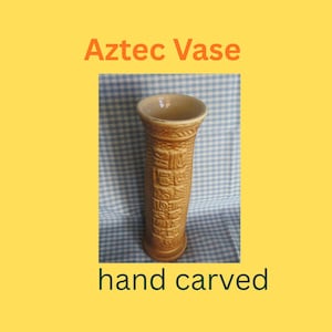 Puede incluir: Un jarrón azteca beige tallado a mano con forma cilíndrica. El jarrón presenta tallas en relieve detalladas y el texto "Aztec Vase" y "hand carved". El fondo es un patrón de cuadros azules y blancos.