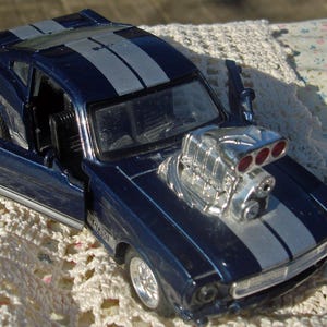 May include: A dark blue toy car with silver racing stripes and a large engine. The car has an open door, revealing the interior. The model car is a GT350.