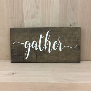 May include: A wooden sign with the word "gather" written in white cursive lettering on a dark brown background.