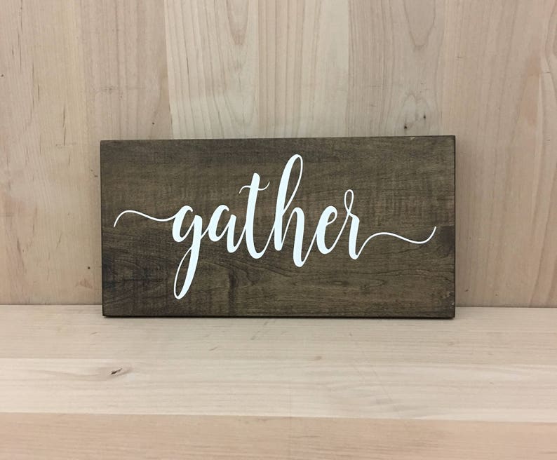 May include: A wooden sign with the word "gather" written in white cursive lettering on a dark brown background.