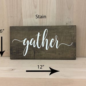 May include: A wooden sign with the word "gather" in white lettering on a dark brown stained background. The sign measures 12 inches wide and 6 inches tall.