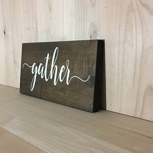 May include: A dark brown wooden sign with the word "gather" written in white cursive script.