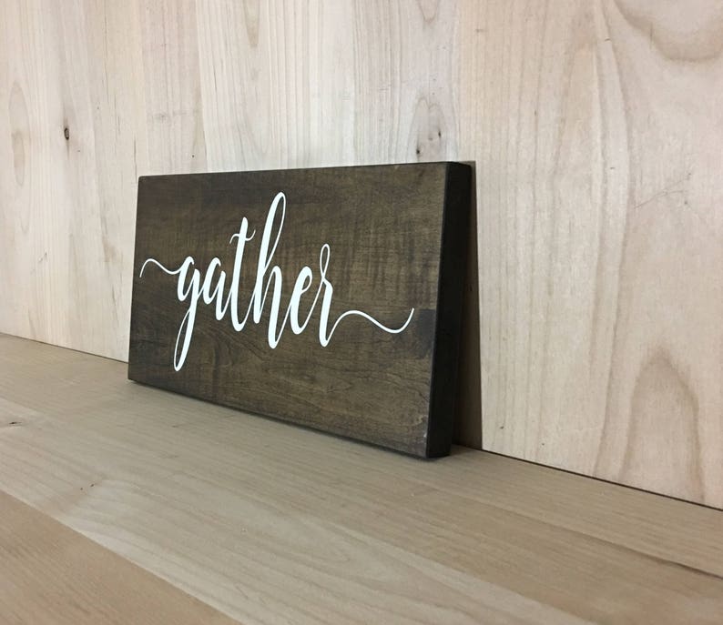 May include: A dark brown wooden sign with the word "gather" written in white cursive script.