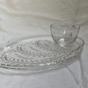 May include: Clear glass teacup and saucer set. The oval-shaped saucer features a decorative leaf pattern. The teacup has a handle and sits on a raised platform on the saucer. The set is made of clear glass.