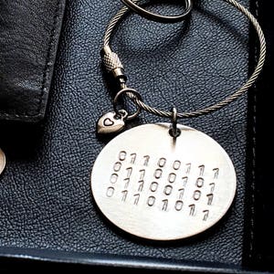 May include: Silver keychain with a round charm engraved with a pattern of 0s and 1s. A small heart charm hangs from the top of the round charm.