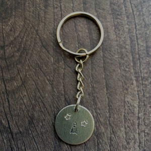 May include: Gold keychain with a round metal tag featuring a rocket ship and stars.