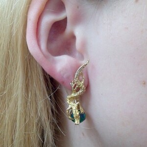 May include: Gold earrings with a green gemstone and a delicate, intricate design. The earrings are shaped like a stylized leaf or branch.