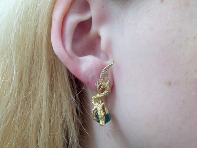 May include: Gold earrings with a green gemstone and a delicate, intricate design. The earrings are shaped like a stylized leaf or branch.