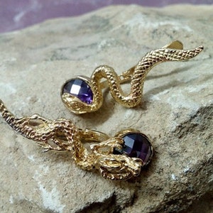 May include: A pair of gold-tone earrings featuring a coiled snake design with a purple gemstone in the center of each earring.