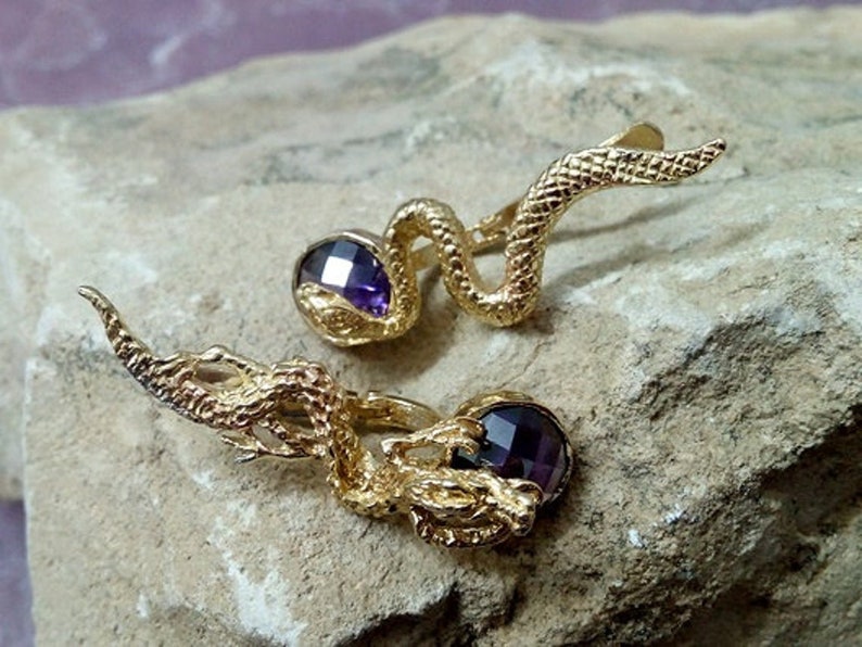 May include: A pair of gold-tone earrings featuring a coiled snake design with a purple gemstone in the center of each earring.