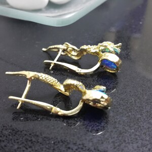 May include: A pair of gold earrings with blue opal stones. The earrings are in the shape of a small, intricate design.