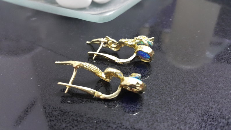 May include: A pair of gold earrings with blue opal stones. The earrings are in the shape of a small, intricate design.