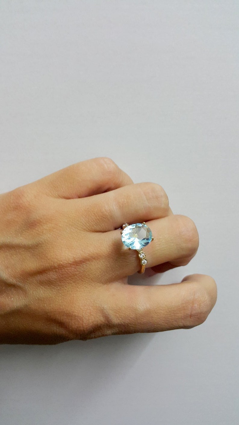 May include: A gold ring featuring a large oval light blue gemstone, with smaller clear gemstones set into the band.