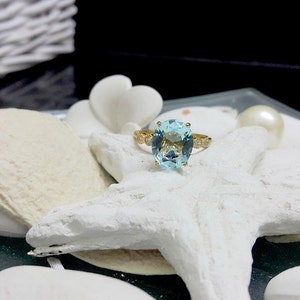 May include: A gold ring featuring a large oval-shaped light blue gemstone, surrounded by smaller clear gemstones. The ring is displayed on a white starfish.