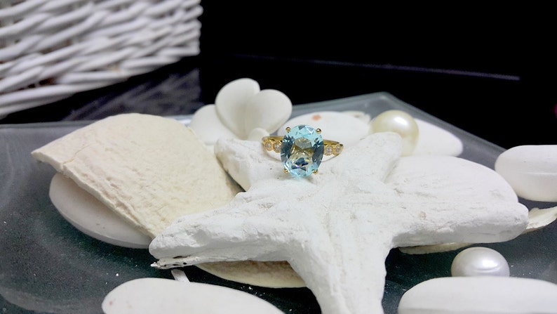 May include: A gold ring featuring a large oval-shaped light blue gemstone, surrounded by smaller clear gemstones. The ring is displayed on a white starfish.
