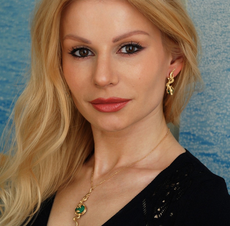 May include: A woman wearing a black top with a gold necklace and earrings. The necklace and earrings feature green stones and gold accents.