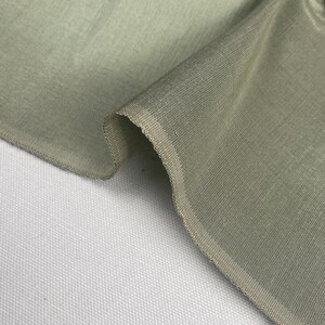 Puede incluir: Primer plano de una sección de tela verde salvia. La tela está doblada, revelando la textura y el tejido. Los bordes son visibles, mostrando la construcción de la tela. El fondo es una superficie blanca texturizada.
