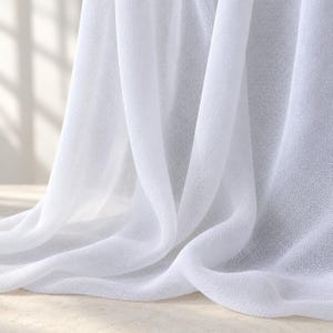 May include: A sheer white curtain panel made of a lightweight, semi-transparent fabric. The material features a fine, woven texture and hangs in soft, vertical folds, allowing light to pass through the translucent surface of the textile.
