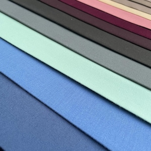 Puede incluir: Una colección de muestras de tela en varios tonos de azul, verde, rosa y tonos neutros. Las telas están dispuestas en filas diagonales, mostrando los diferentes colores y texturas. La imagen está bien iluminada, resaltando las sutiles variaciones de color y material.