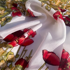 Puede incluir: Pañuelo de seda blanco con un vibrante estampado floral. El diseño presenta grandes amapolas rojas con tallos y hojas verdes, sobre un fondo de follaje amarillo y verde. El pañuelo está drapeado, mostrando su textura fluida.