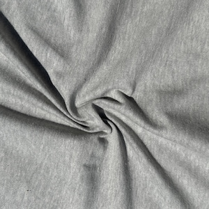 Pode incluir: Close-up de um tecido cinza com uma aparência macia e texturizada. O tecido é dobrado e drapeado, criando sombras e destaques. O material parece ser um tecido de malha ou jersey, adequado para roupas ou decoração de casa.