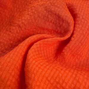 Pode incluir: Close-up de um tecido laranja vibrante com uma trama texturizada. O tecido está dobrado, criando sombras e destacando a textura do material. A cor brilhante sugere uma peça de roupa de verão ou um artigo de decoração.