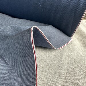 Puede incluir: Primer plano de un rollo de tela vaquera. La tela es de color azul medio con una textura sutil. El borde de la tela está acabado con una raya blanca y roja. La tela está doblada, revelando el reverso.