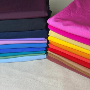 Puede incluir: Montones de tela colorida en varios tonos, incluyendo morado, negro, azul y colores vibrantes como rosa, amarillo y rojo. La tela parece estar doblada cuidadosamente, mostrando una gama de colores adecuados para proyectos de costura o manualidades.