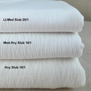 Puede incluir: Se muestran tres pilas de tela blanca, cada una con una textura diferente. La capa superior está etiquetada como "Lt.Med Slub 20/1", la capa intermedia como "Med.Hvy Slub 16/1" y la capa inferior como "Hvy Slub 10/1".