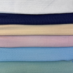 Puede incluir: Primer plano de una pila de muestras de tela en varios colores. Los colores incluyen blanco, azul marino, beige, rosa claro, azul claro y verde espuma de mar. Cada tela tiene un sutil patrón texturizado.