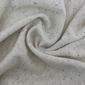 Puede incluir: Primer plano de una tela de punto color crema con pequeñas motas azules dispersas. La tela está doblada, mostrando la textura y la caída del material. El tejido parece suave y posiblemente adecuado para ropa.