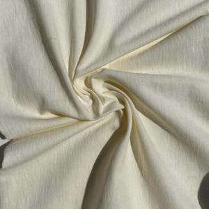 Pode incluir: Close-up de um tecido creme com dobras e sombras suaves. O tecido parece ser um material leve, possivelmente algodão ou linho, com um brilho sutil. A textura é lisa e a aparência geral é elegante.