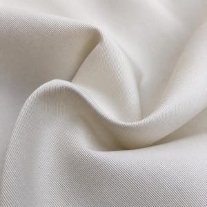 May include: A close-up view of off-white fabric featuring a distinct diagonal twill weave pattern. The material is folded into soft, flowing curves, highlighting the texture and the consistent, tight weave of the textile surface throughout.