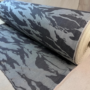 Puede incluir: Un rollo de tela con un patrón de camuflaje en tonos grises y azules. La tela está enrollada, revelando los detalles del patrón. El diseño presenta formas irregulares, creando una apariencia texturizada. Adecuado para ropa y decoración del hogar.