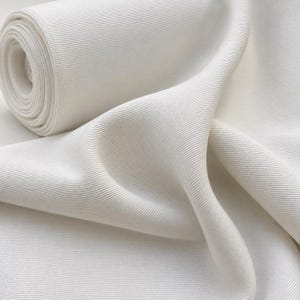 May include: A roll of off-white fabric featuring a distinct diagonal twill weave pattern. The material is shown partially unrolled, creating soft, flowing folds that highlight the textured surface and the consistent, dense weave of the textile.