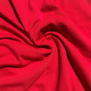 Pode incluir: Close-up de um tecido vermelho vibrante com uma textura macia e fluida. O material é tecido de forma apertada, criando uma superfície lisa com sombras e reflexos sutis. O tecido é reunido em espiral, mostrando sua queda.